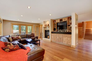 Ein großzügiges Wohnzimmer mit einer Ledersofa, einem Couchtisch und einem Kamin. Ein Fernseher ist an einem Holzkasten befestigt und eine Treppe führt zu einer oberen Ebene.