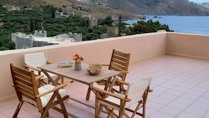 Une terrasse avec une table et des chaises donnant sur un paysage côtier, avec des collines et des bâtiments.