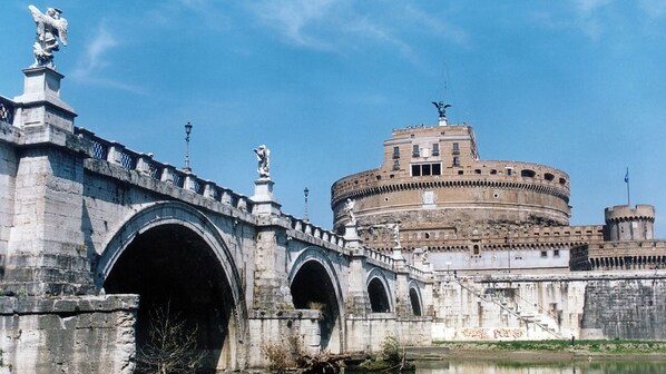 A stone bridge with arches, a statue, and a large historic building with a dome in the background.