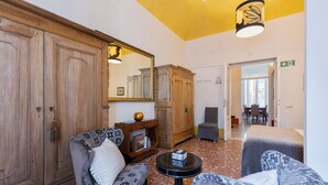 A room with a wooden wardrobe, two patterned armchairs, a small round table with a book, and a mirror.