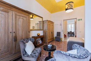 A room with a wooden wardrobe, two patterned armchairs, a small round table with a book, and a mirror.