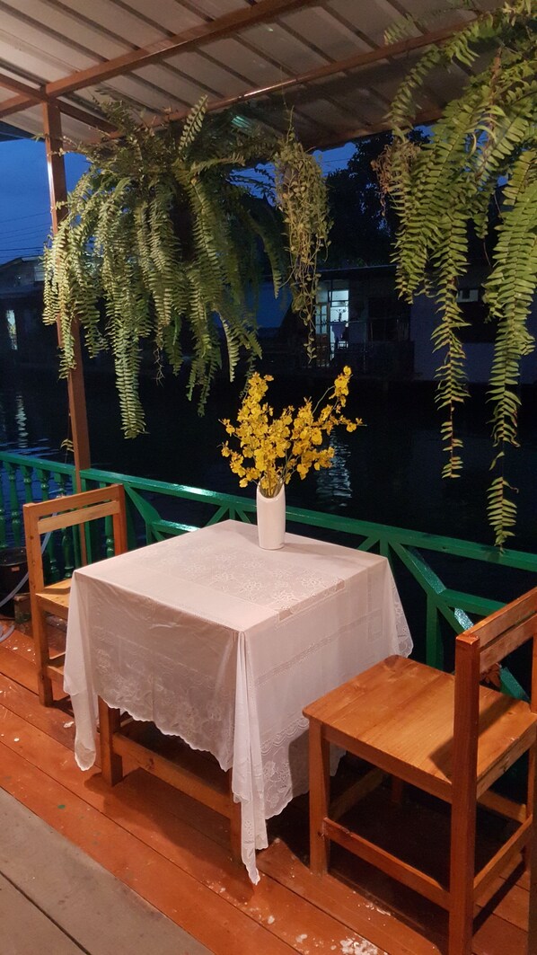 Una mesa exterior con mantel blanco, dos sillas de madera y un jarrón con flores amarillas.