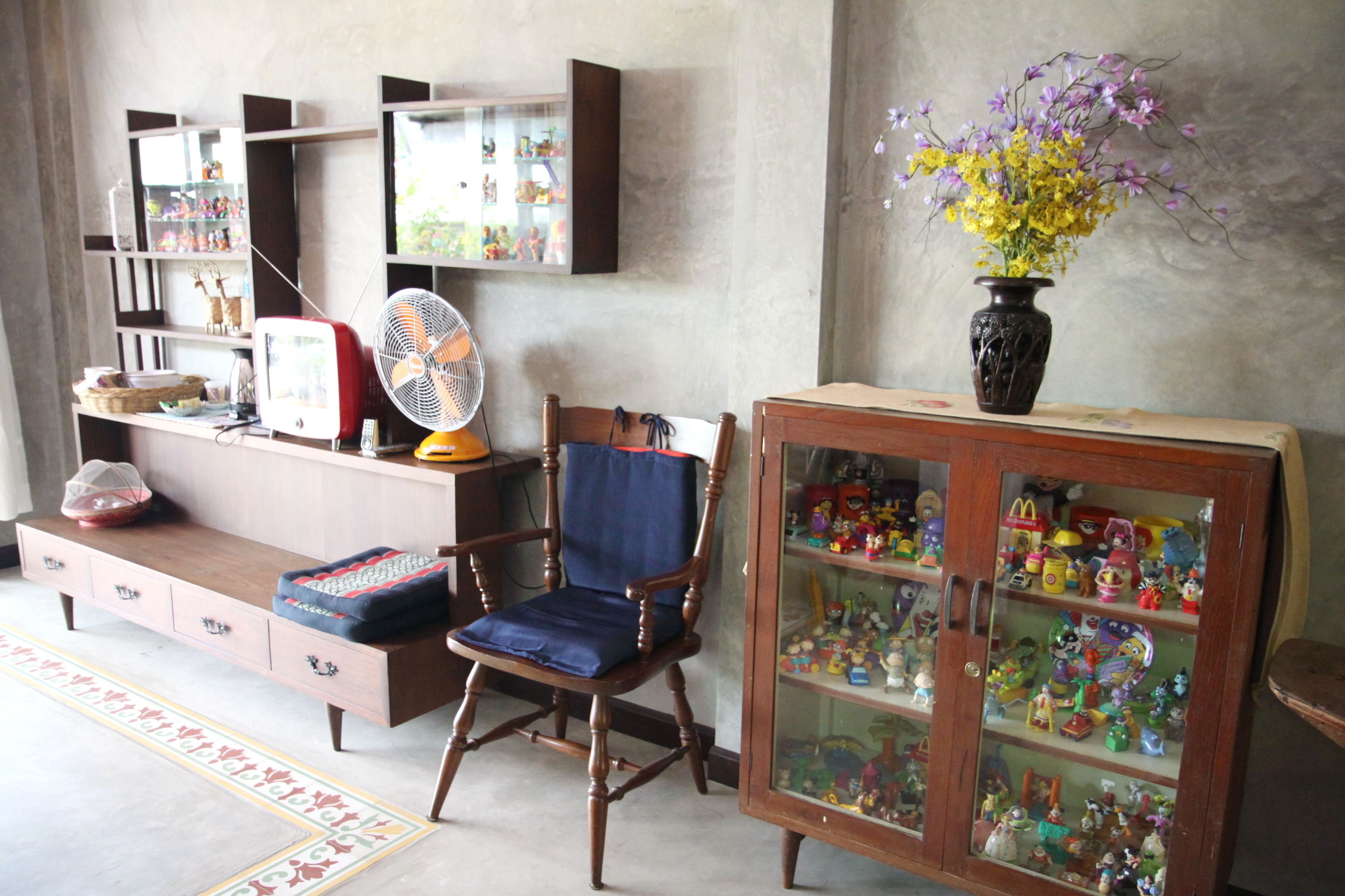 A room with a wooden cabinet filled with toys, a blue chair, a fan, and a vase with flowers.