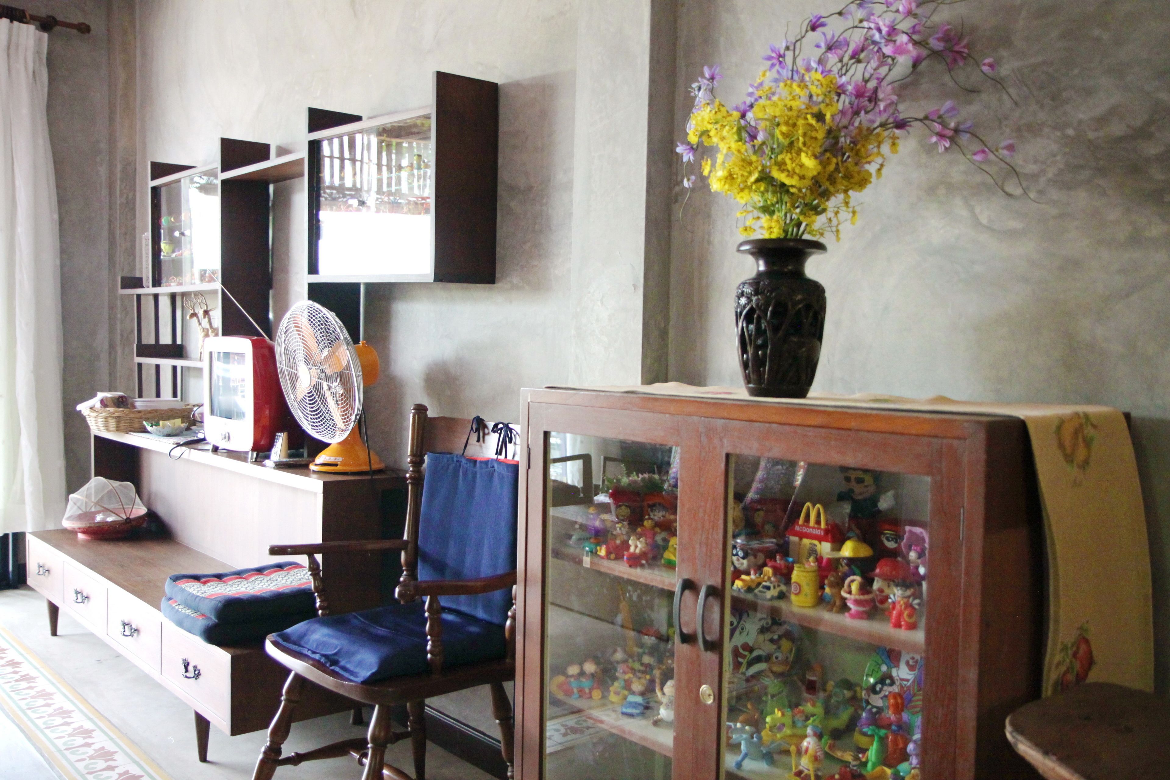 A living room with a wooden cabinet displaying collectibles, a blue cushioned chair, a fan, and a vase with flowers.