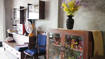 A living room with a wooden cabinet displaying collectibles, a blue cushioned chair, a fan, and a vase with flowers.