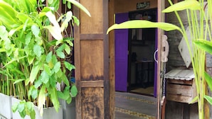 Un porche de madera con dos macetas de terracota con plantas, una puerta de madera y un interior morado visible a través de la abertura.
