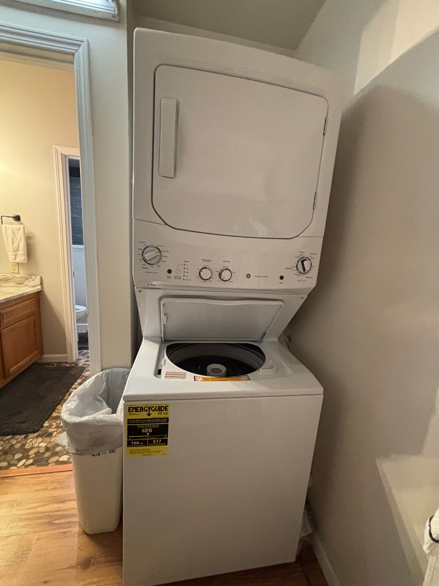 A compact, white front-loading washer and dryer unit with a ENERGY GUIDE sticker, located in a small laundry room with a wooden floor and a door leading to another room.