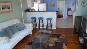 Un salon avec un canapé blanc, une table basse en verre et un tapis à motifs. On y trouve trois tabourets de bar bleus et un coin cuisine avec un réfrigérateur blanc et un évier.