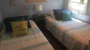 Une chambre avec deux lits, une table de chevet, une lampe, un tableau encadré au mur et une fenêtre avec des rideaux.