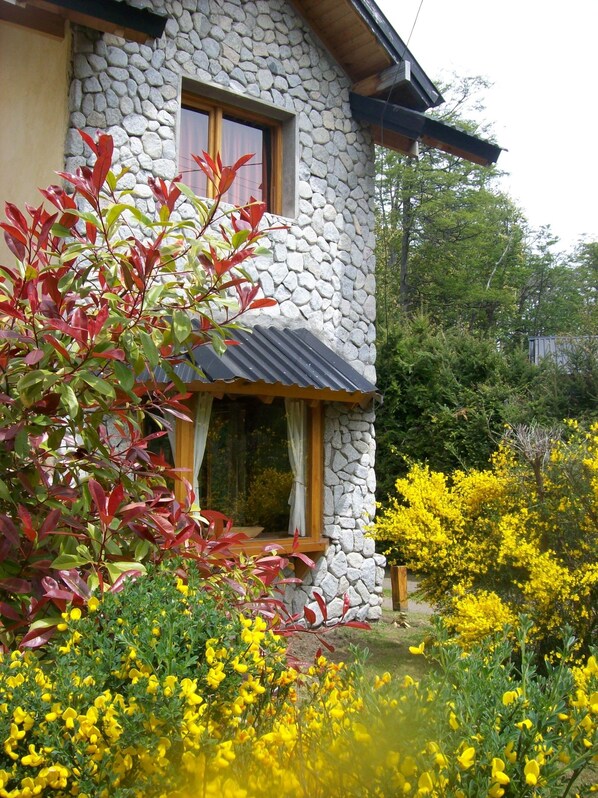 A stone house with a window and a door, surrounded by yellow flowers and red foliage. - Tierras del Sur (Villa La Angostura)