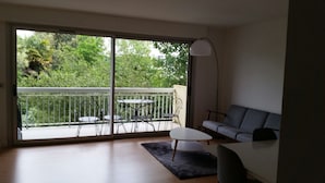 A modern living room with a grey sofa, a small round table, and a floor lamp. Large sliding glass doors lead to a balcony with a white railing and two chairs. - T3 - Quartier Le Parc 200 m from the beach, parking, elevator, terrace, shops (Royan)