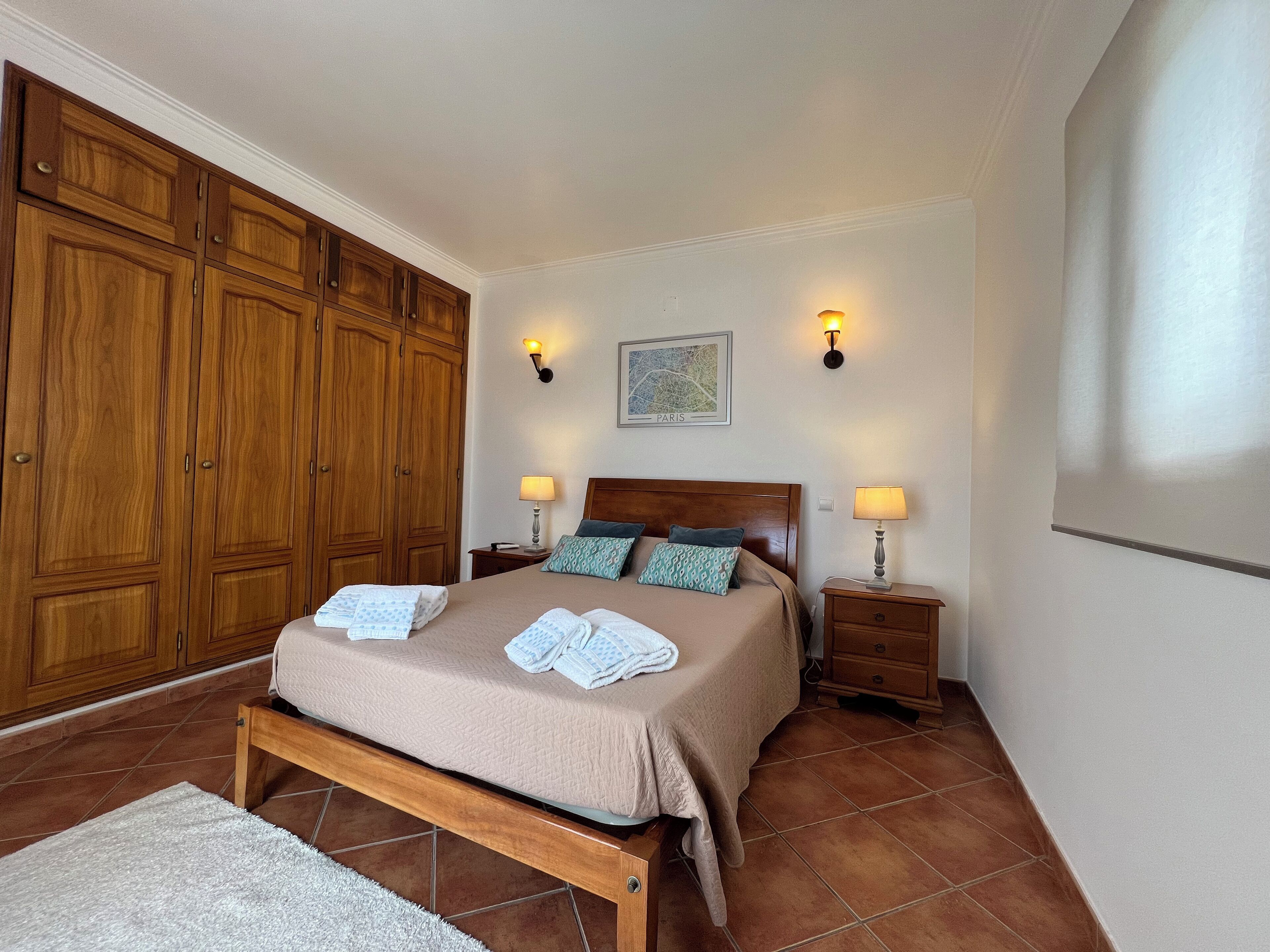 Un dormitorio con un amplio armario de madera, una cama con cubrecama color beige, dos lámparas de noche y un mapa de París colgado en la pared.