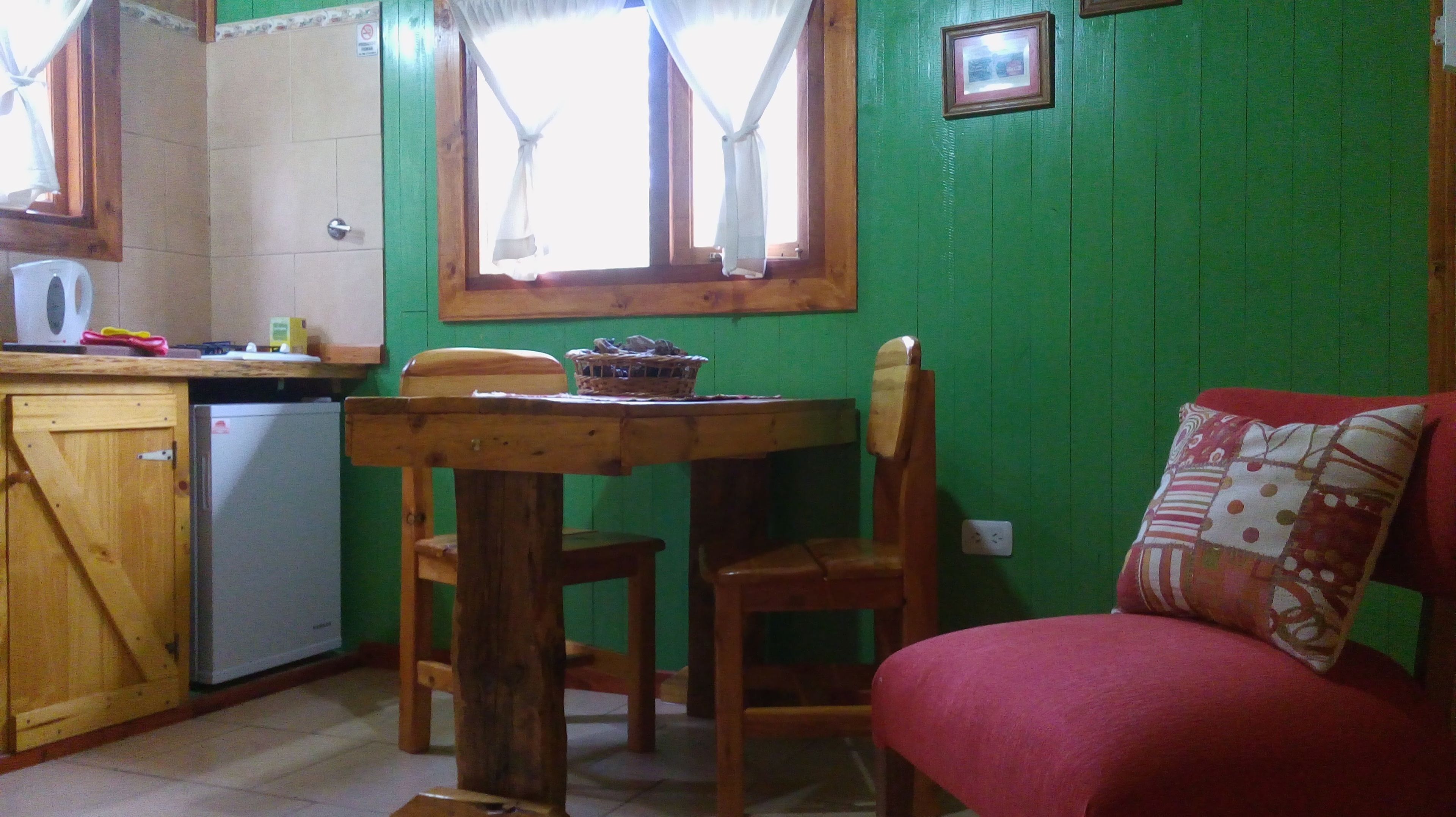 A compact kitchen with a wooden table and chairs, a small refrigerator, and a red sofa with a patterned pillow.