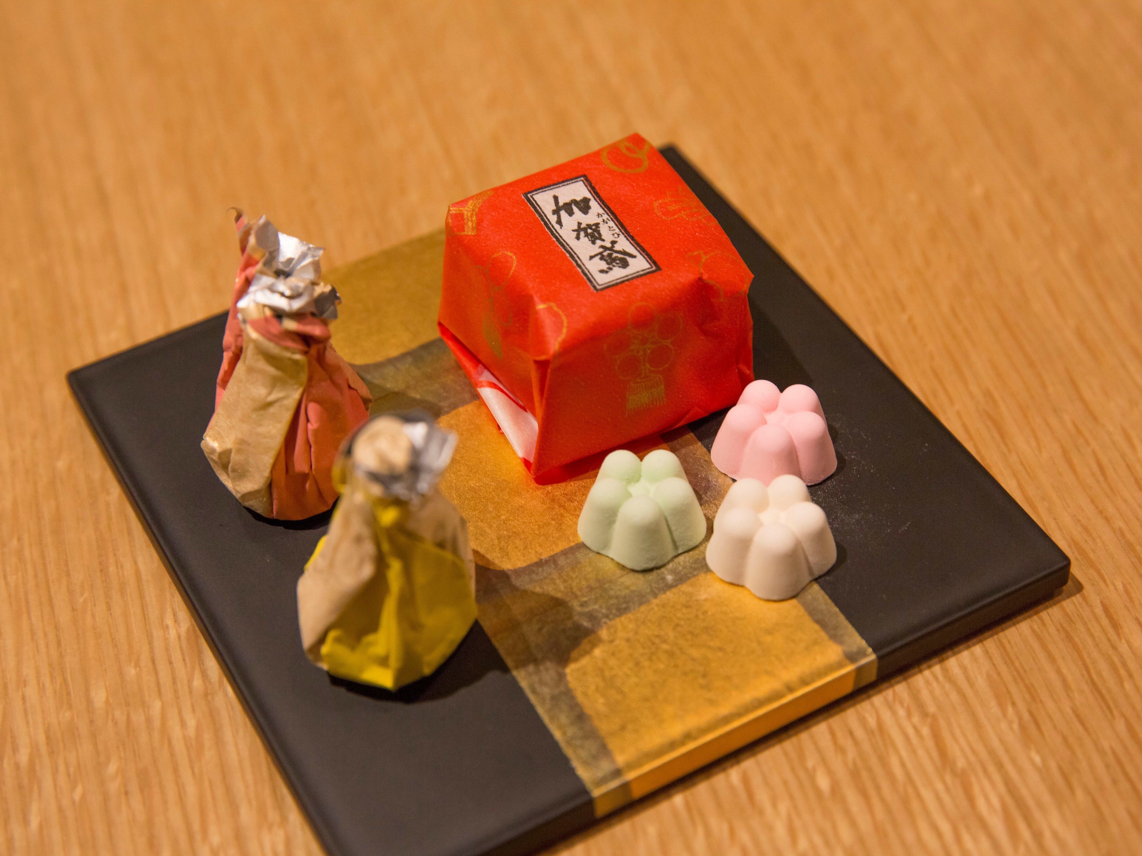 A black tray with a red block, a yellow wrapped item, and three pink and white items on a wooden surface.