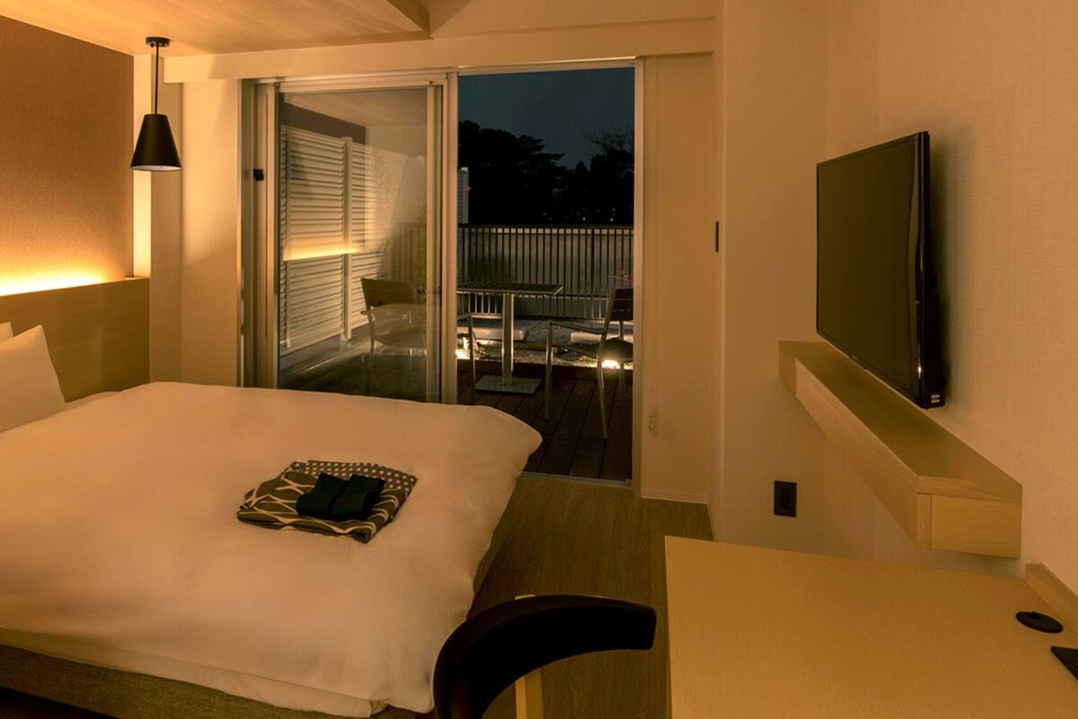 Habitación de hotel con cama, escritorio, televisor, balcón con asientos exteriores y puerta corrediza.