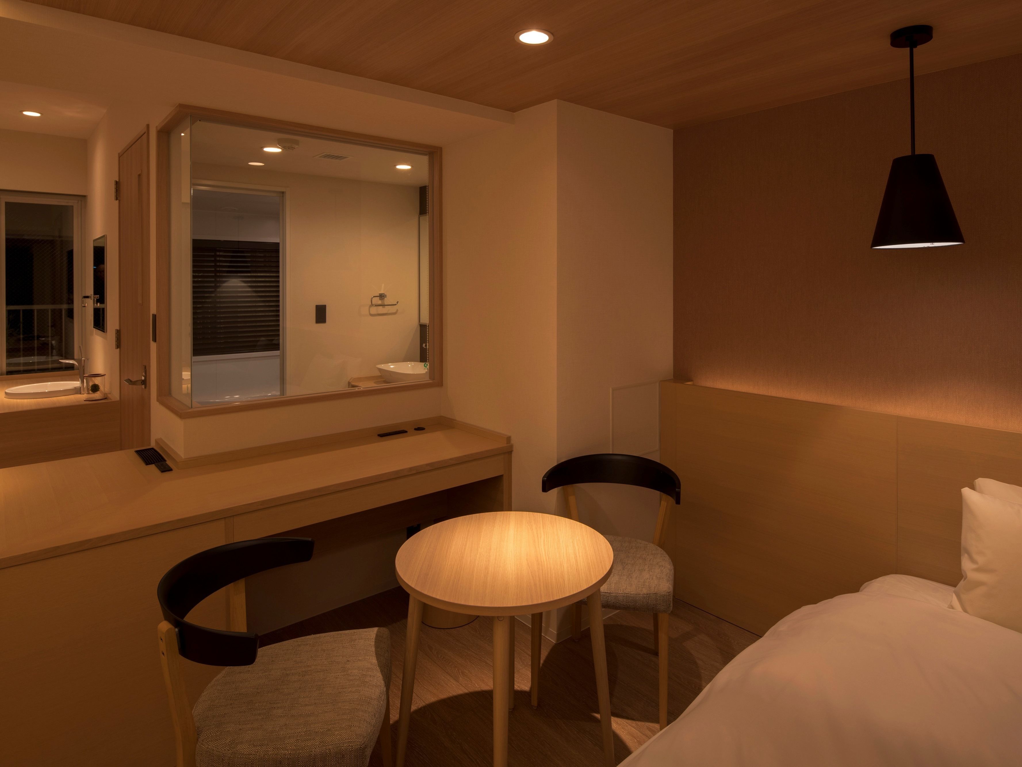 A modern hotel room with a large mirror, a round table, two chairs, a bed, and a hanging light fixture.