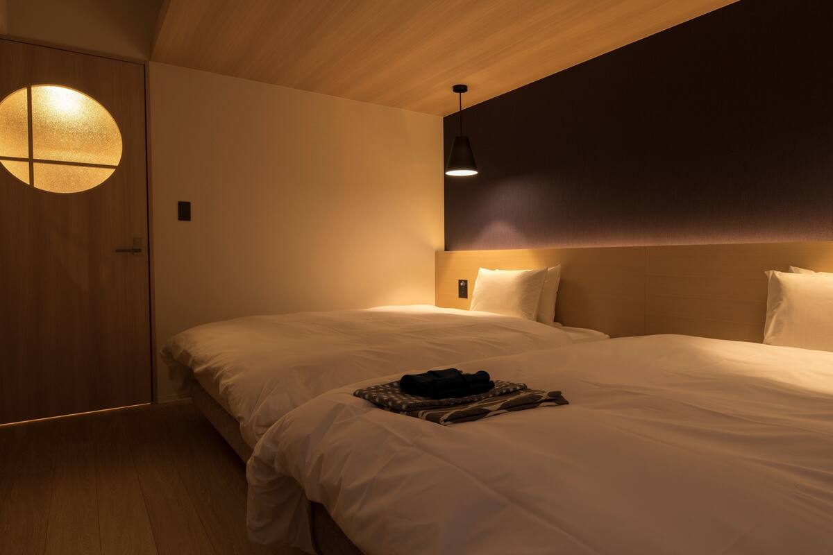 Habitación de hotel con techo de madera, una lámpara de pared redonda, cama con sábanas blancas y cabecero oscuro.