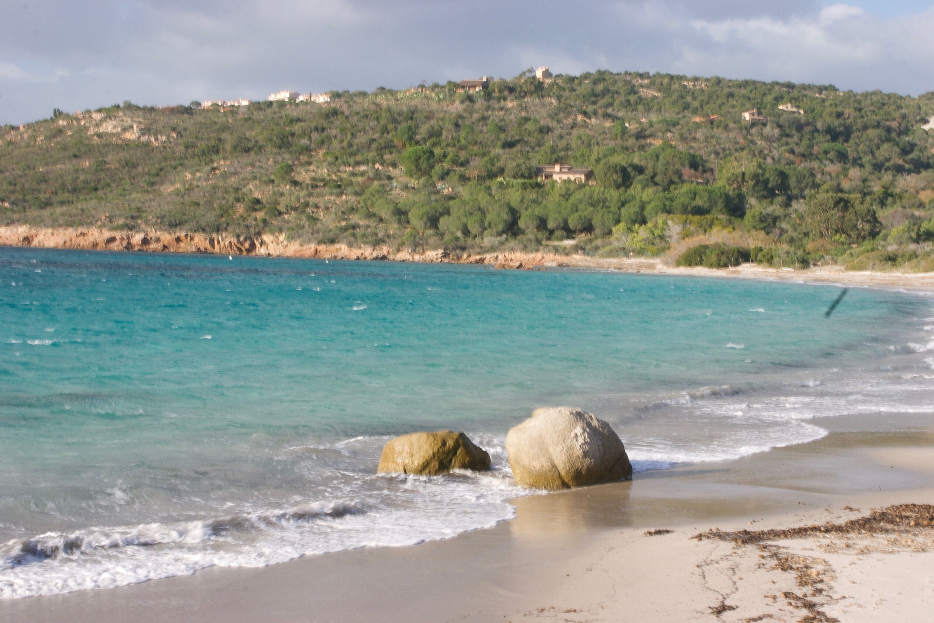 Ein Strand mit klarem türkisblauem Wasser, einem großen Felsen im Vordergrund und einer Hügelkuppe mit Gebäuden im Hintergrund.
