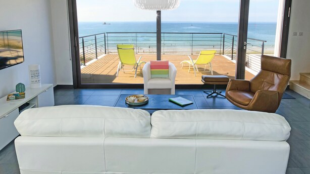 A modern living room with a large sofa, a dining table with chairs, and a view of the ocean through a sliding glass door. - Beachfront villa, spectacular sea view, electric charging station (Plouescat)