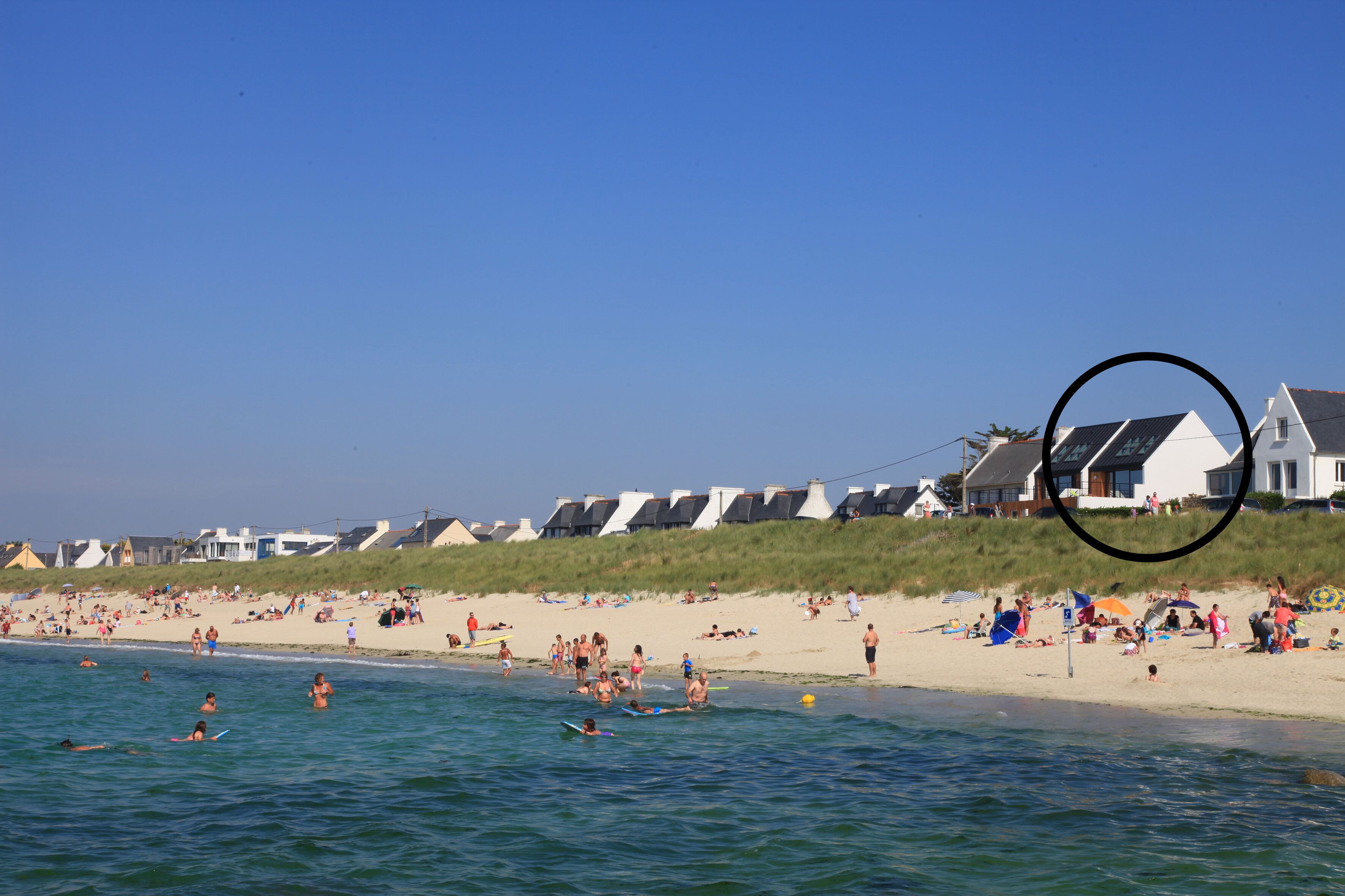 Une plage où l’on voit des gens nager et faire de la planche à voile, un rivage sablonneux et des maisons en arrière-plan.