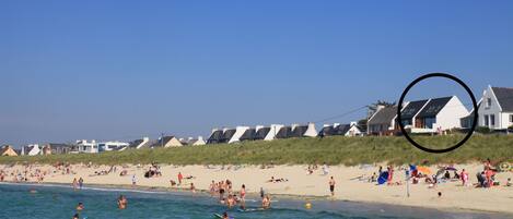 Une plage où l’on voit des gens nager et faire de la planche à voile, un rivage sablonneux et des maisons en arrière-plan.