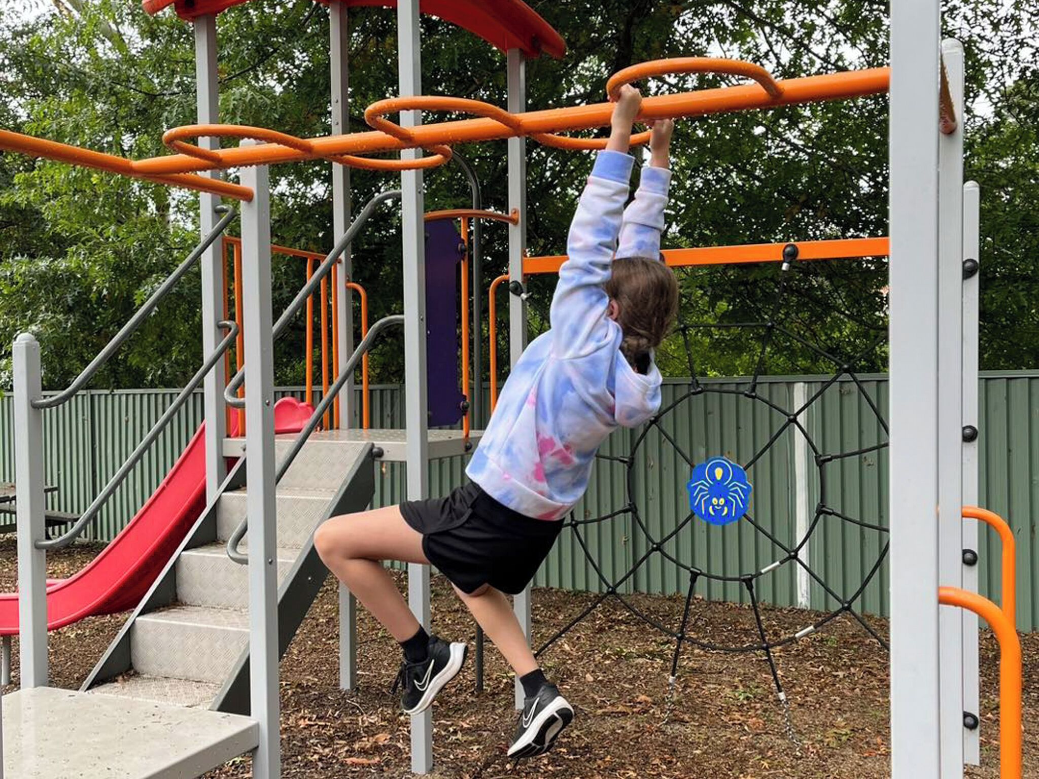A child climbing monkey bars at a playground with a red slide and a spider-themed sign.