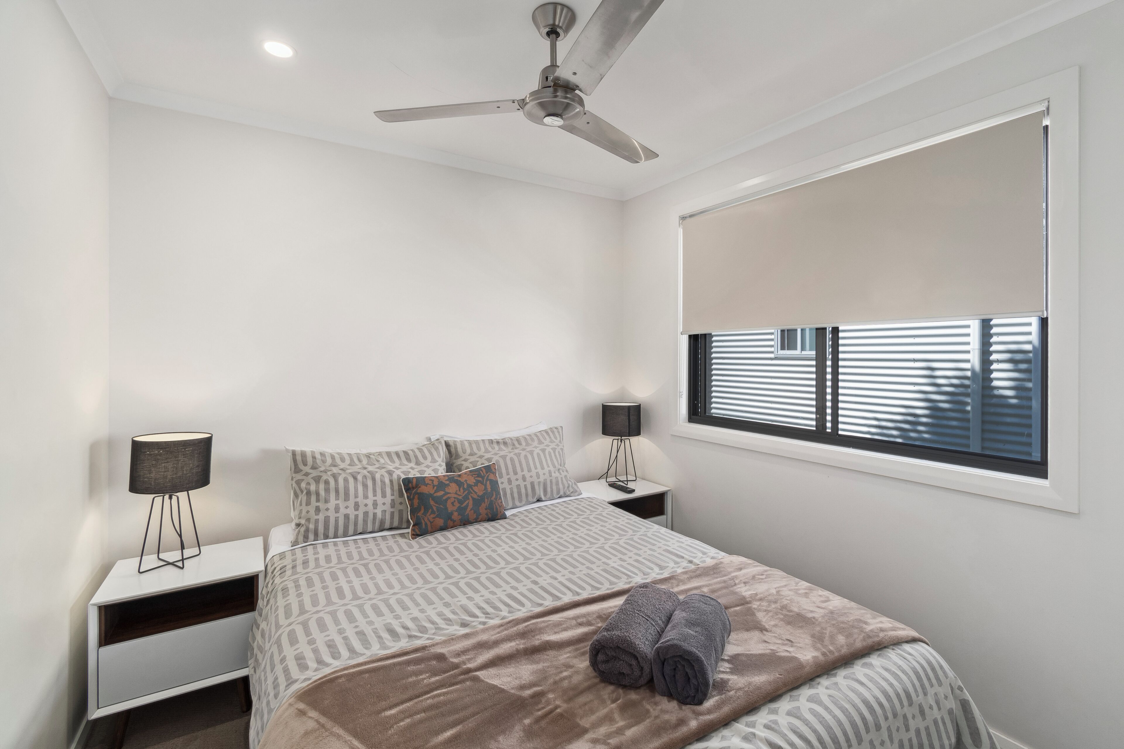 A bedroom with a bed, two bedside tables, a ceiling fan, a window with blinds, and a nightstand with a lamp.