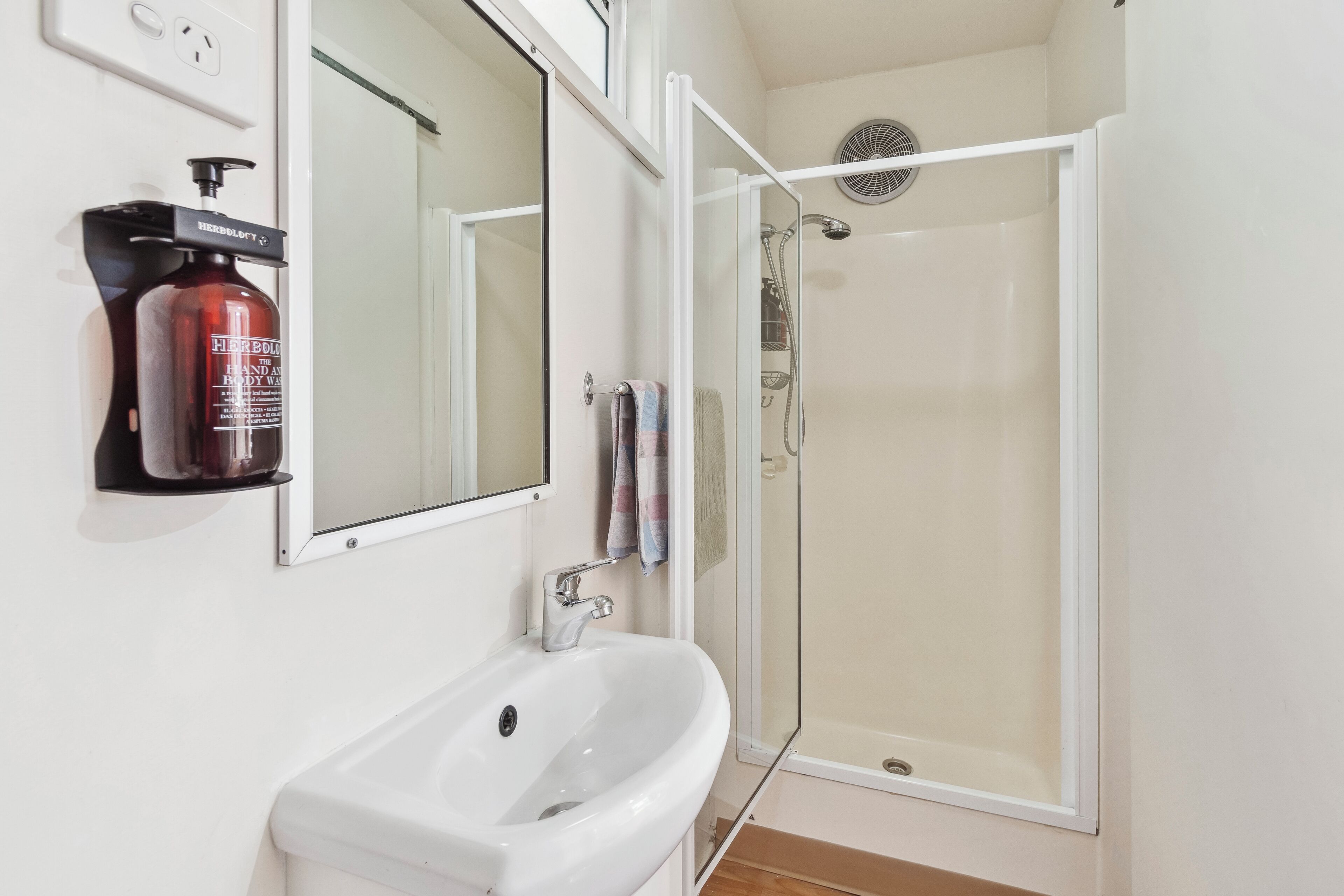 A bathroom with a white sink, a mirror, a shower with a glass door, and a bottle of body lotion on the wall.