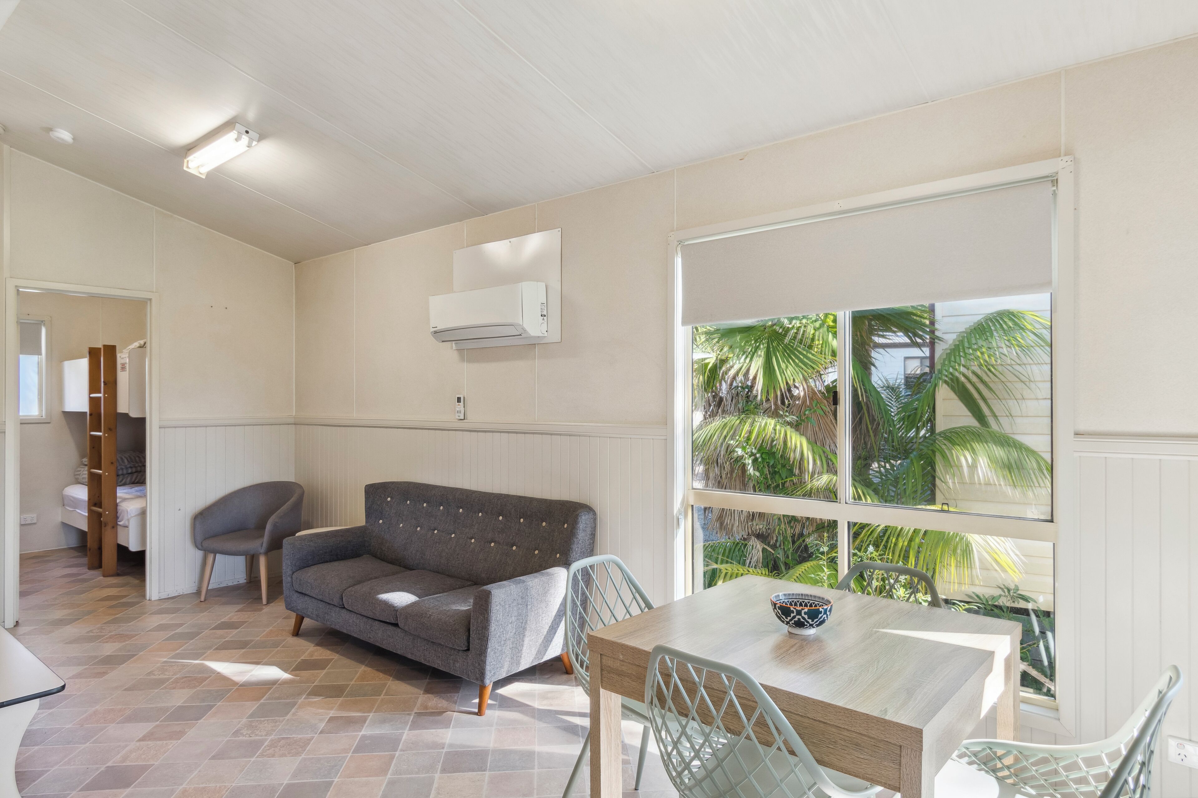 A room with a wooden dining table, a grey sofa, and a window with a view of palm trees.