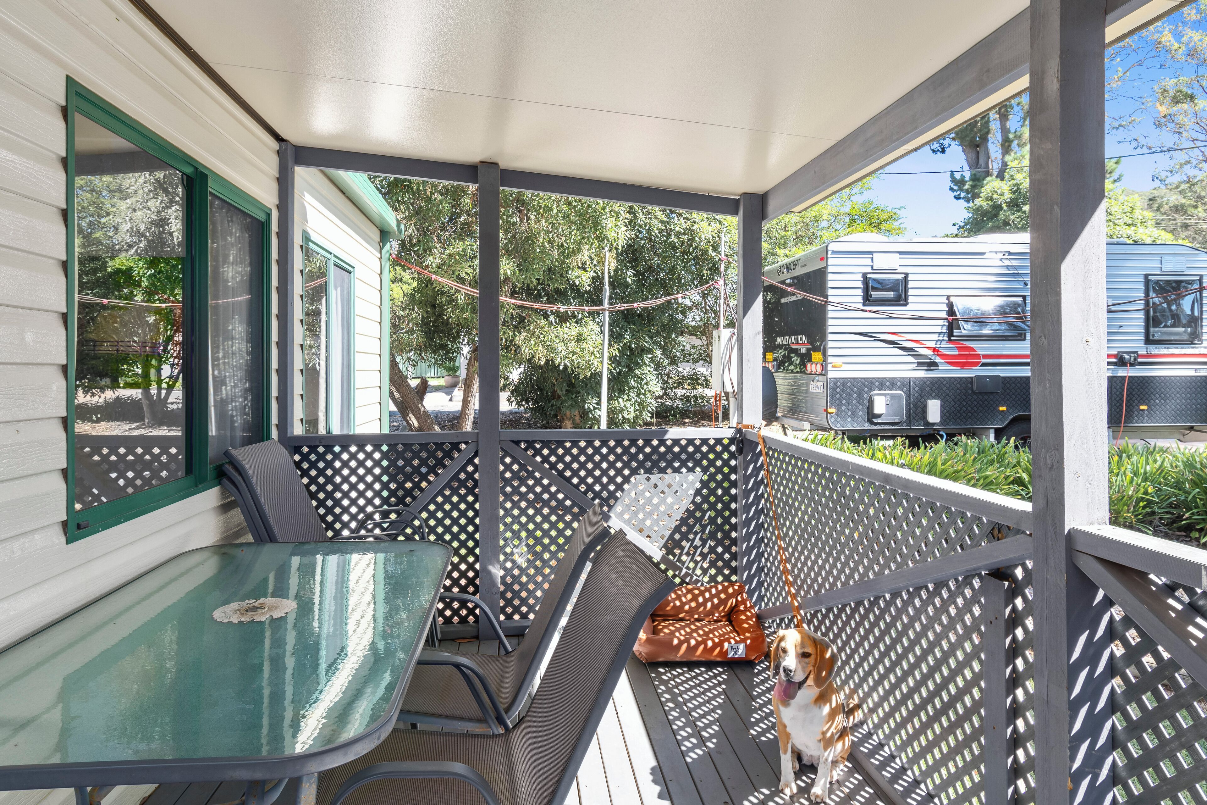 A covered porch with a table and chairs, a dog bed, and a view of a trailer park with a trailer.