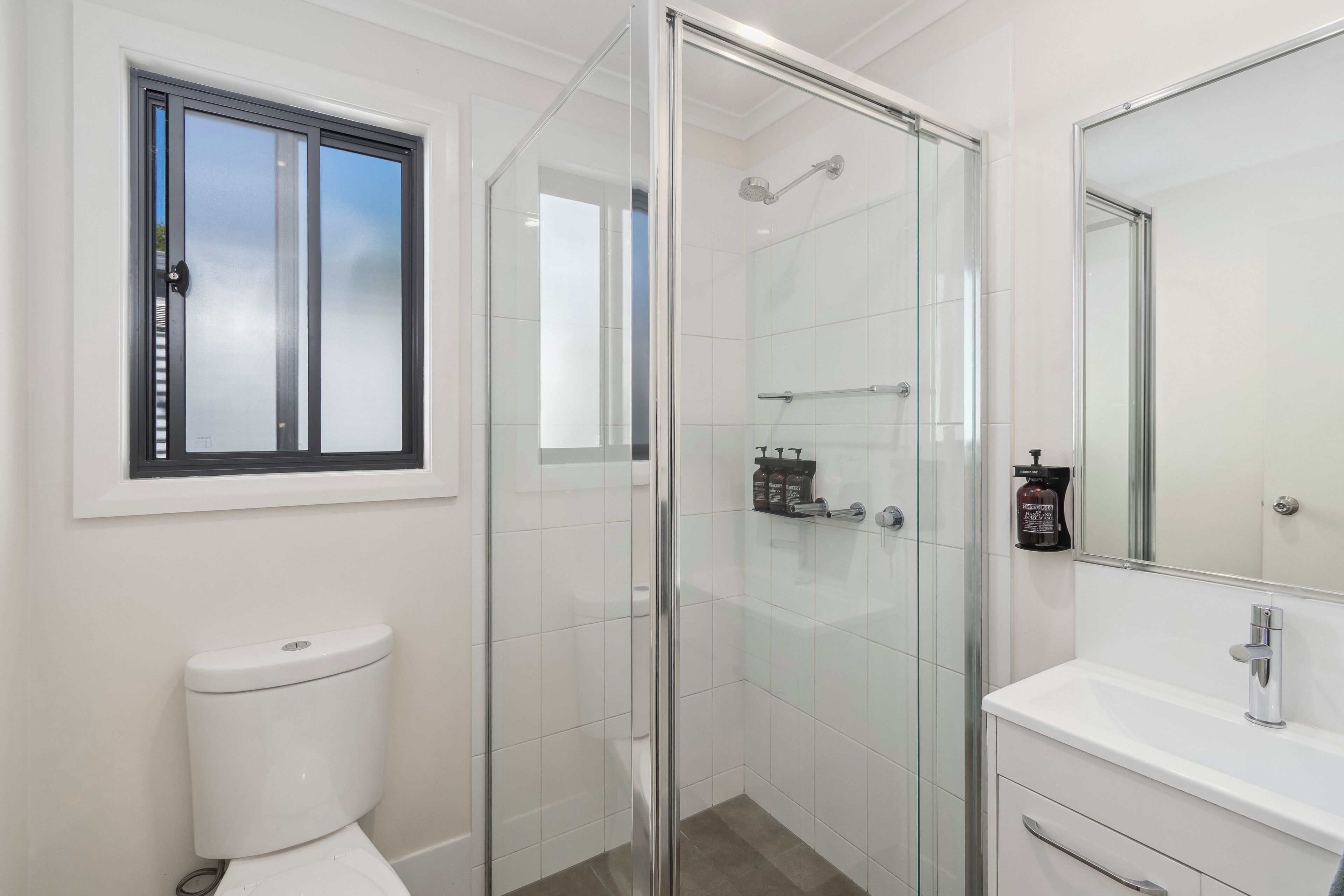 A modern bathroom with a glass-enclosed shower, a white toilet, a sink with a mirror, and a window above the toilet.