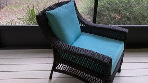 A wicker chair with a teal cushion on a wooden deck, overlooking a garden with a bench and a bird feeder.