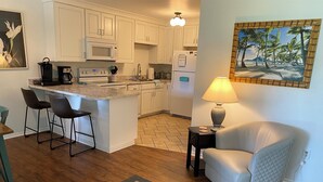 A modern kitchen with white cabinets, a central island with bar stools, a refrigerator, and a microwave. There's a dining area with a table and chairs, a lamp, and a painting of a beach scene.