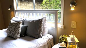Un dormitorio con cama, almohadas, mesita de noche con una planta y ventana con vistas a árboles.