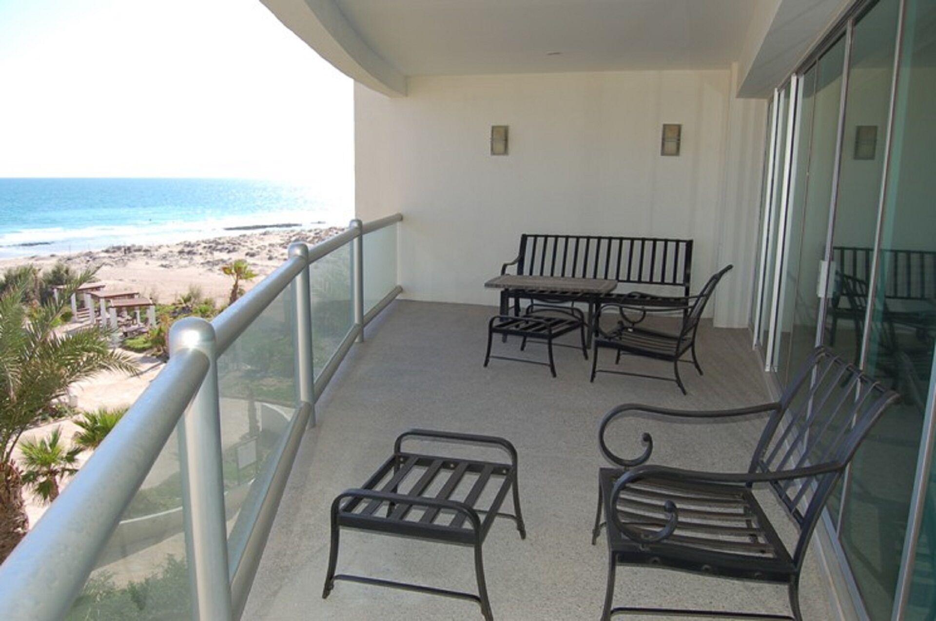 A balcony with a view of the beach, a table, two chairs, and a bench.