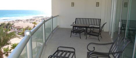 A balcony with a view of the beach, a table, two chairs, and a bench.