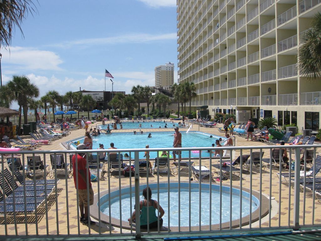 Un hotel con una grande piscina esterna circondata da lettini e ombrelloni, un edificio alto con balconi e una palma.