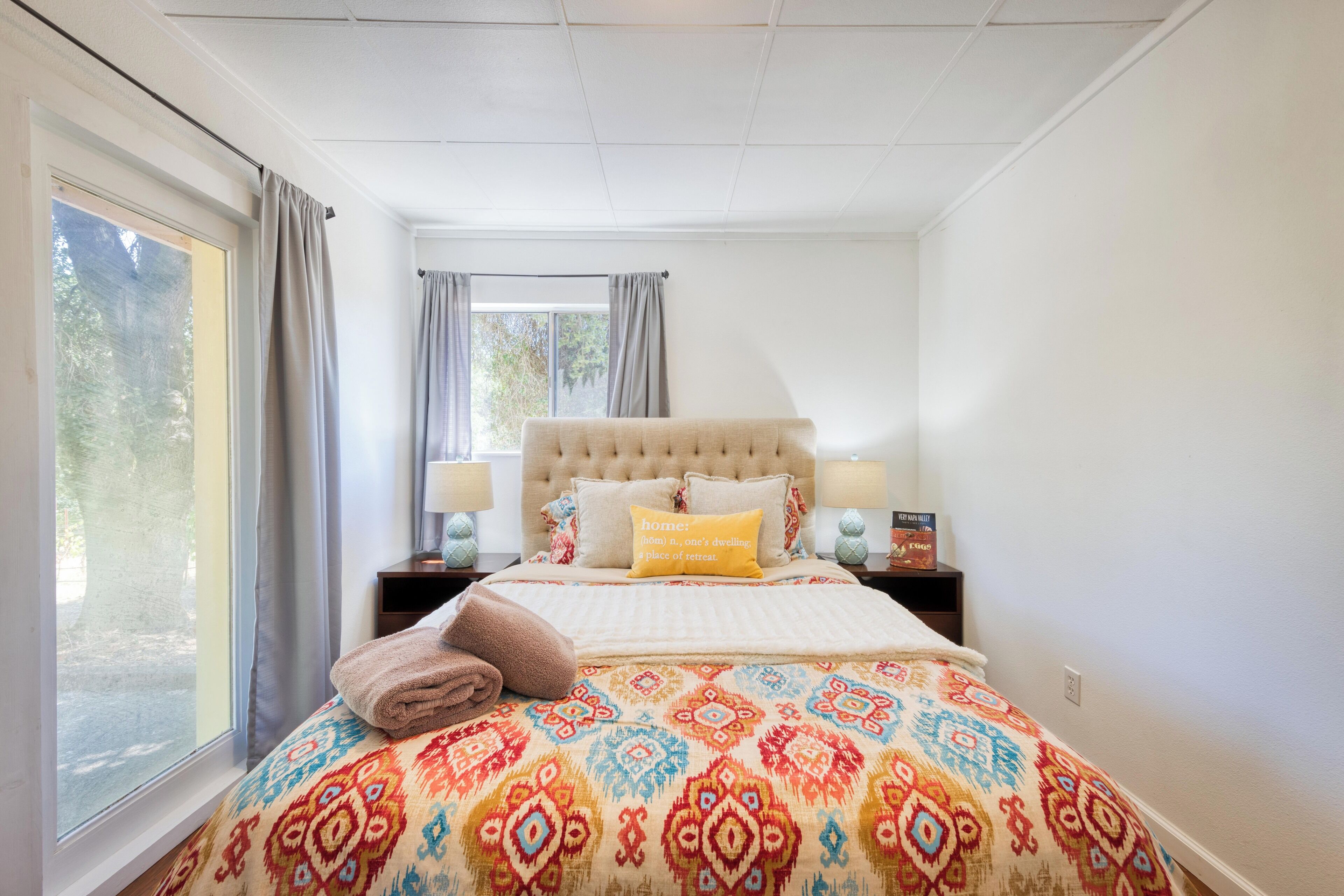A bedroom with a large bed, a window with curtains, a nightstand with a lamp, and a bedside table with a book.