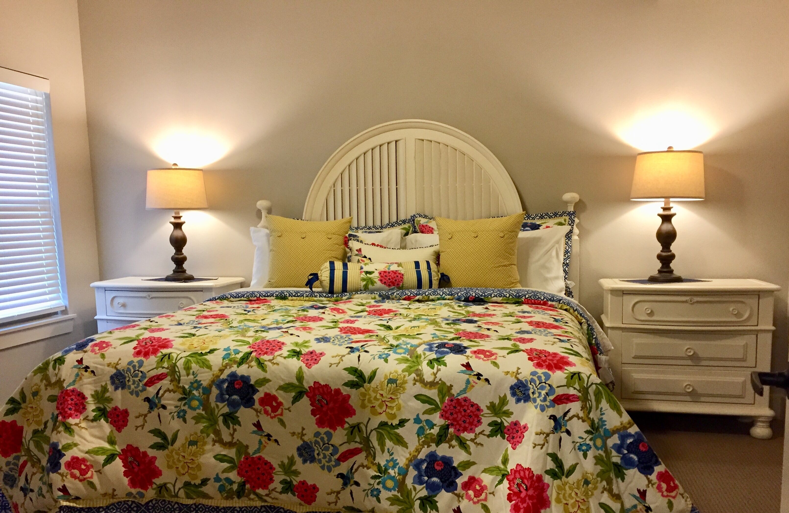 A bedroom with a floral bedspread, two bedside lamps, a white headboard, and a nightstand with drawers.