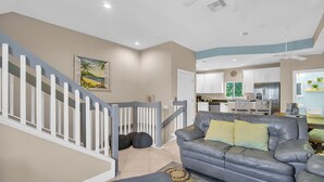 A modern living room with a gray leather sofa, a staircase with white railings, and a kitchen in the background.