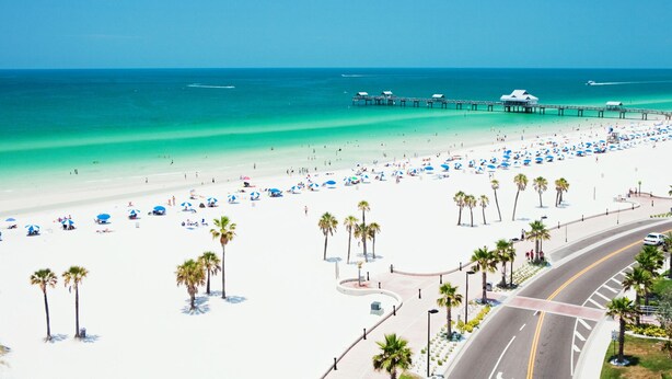 A beach with a pier, palm trees, and a road curving around the beach.