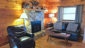 Interior acogedor de una cabaña de troncos con chimenea de piedra, un sillón de cuero y un sofá con cojines estampados.