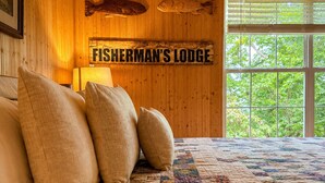 Uma cama com almofadas, um edredom e um letreiro com a inscrição 'FISHERMAN'S LODGE' em um quarto com paredes de madeira e uma janela.