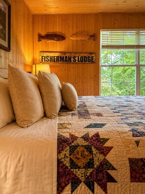 Un letto con cuscini, una coperta e un cartello con su scritto 'FISHERMAN'S LODGE' in una stanza dalle pareti in legno e una finestra.