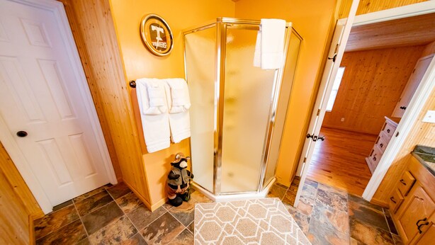 A bathroom with a glass shower, wooden walls, a white door, and a patterned rug.