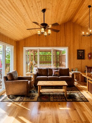 Ein geräumiges Wohnzimmer mit Holzboden, Deckenventilator, Kamin und großem Fenster mit Blick auf eine Terrasse.