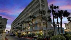 Un hotel de varios pisos con balcones, palmeras y una zona de estacionamiento.