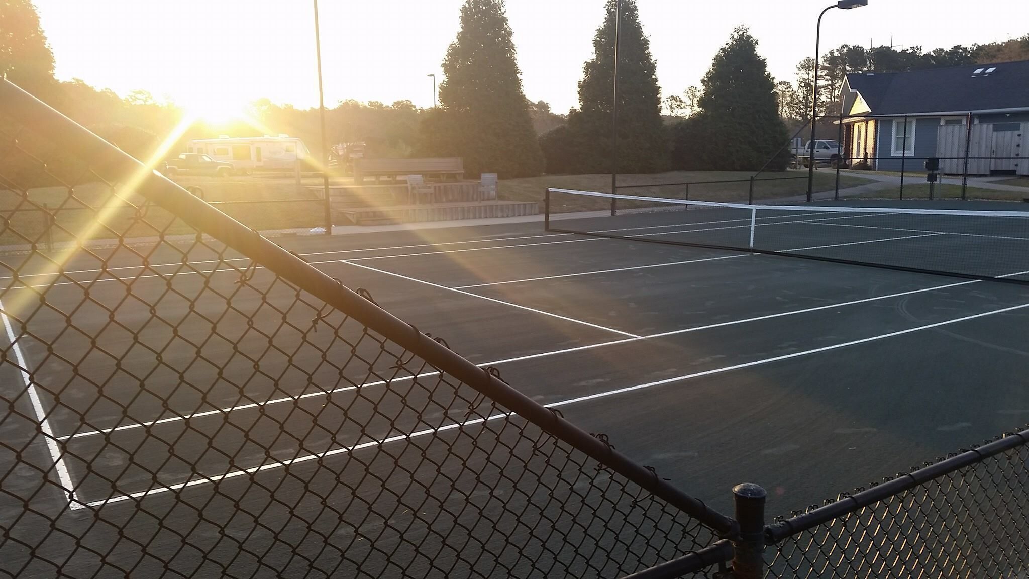 Un court de tennis en plein air, entouré d’une clôture en treillis métallique, bordé d’arbres et avec un bâtiment visible au loin.