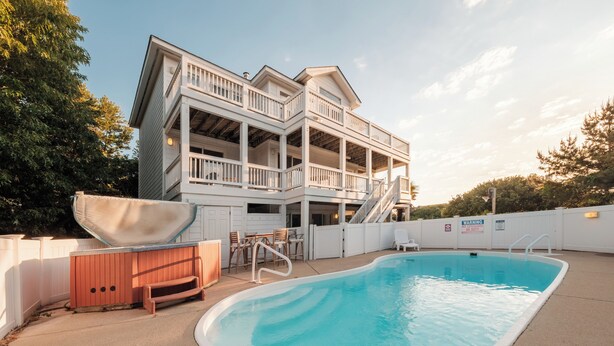 A two-story house with a swimming pool, outdoor furniture, and a wooden bench. - DolphinInnOBX Steps to beach 7BR Pool, Gym, Pickleball/Tennis, Pirate Ship, Golf (Southern Shores)