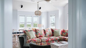 A modern living room with a sectional sofa, a ceiling fan, and large windows. - DolphinInnOBX Steps to beach 7BR Pool, Gym, Pickleball/Tennis, Pirate Ship, Golf (Southern Shores)
